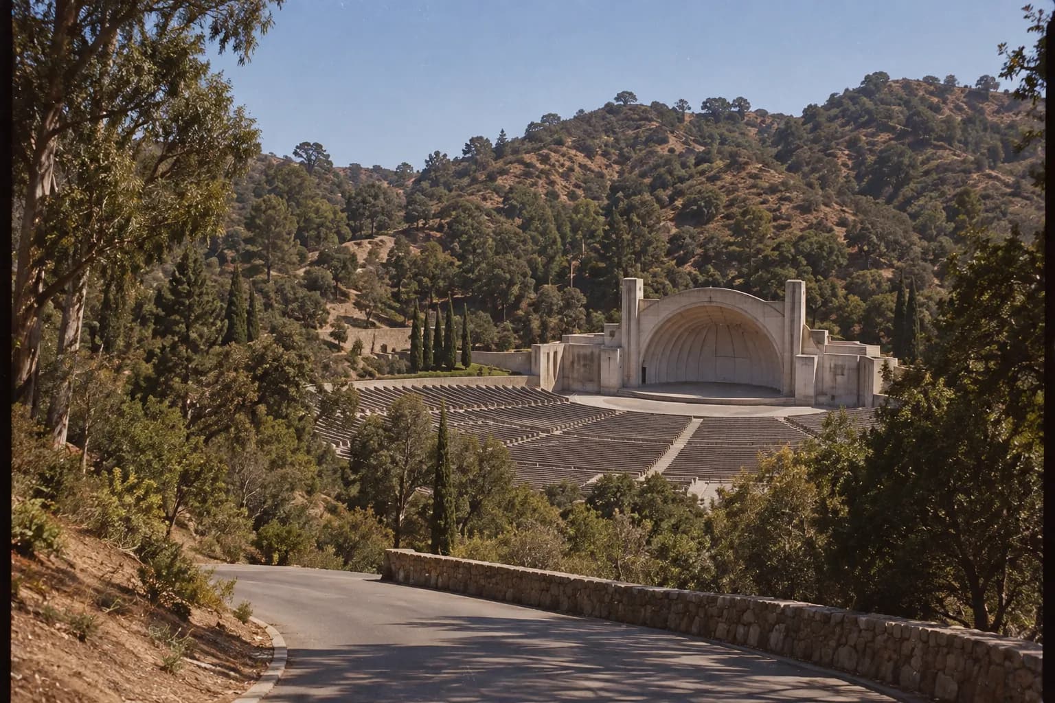 Wide landscape photograph of an open-air theatre set within a Los Angeles County regional park, showing rows of seats, the concrete stage structure and the surrounding Hollywood Hills.