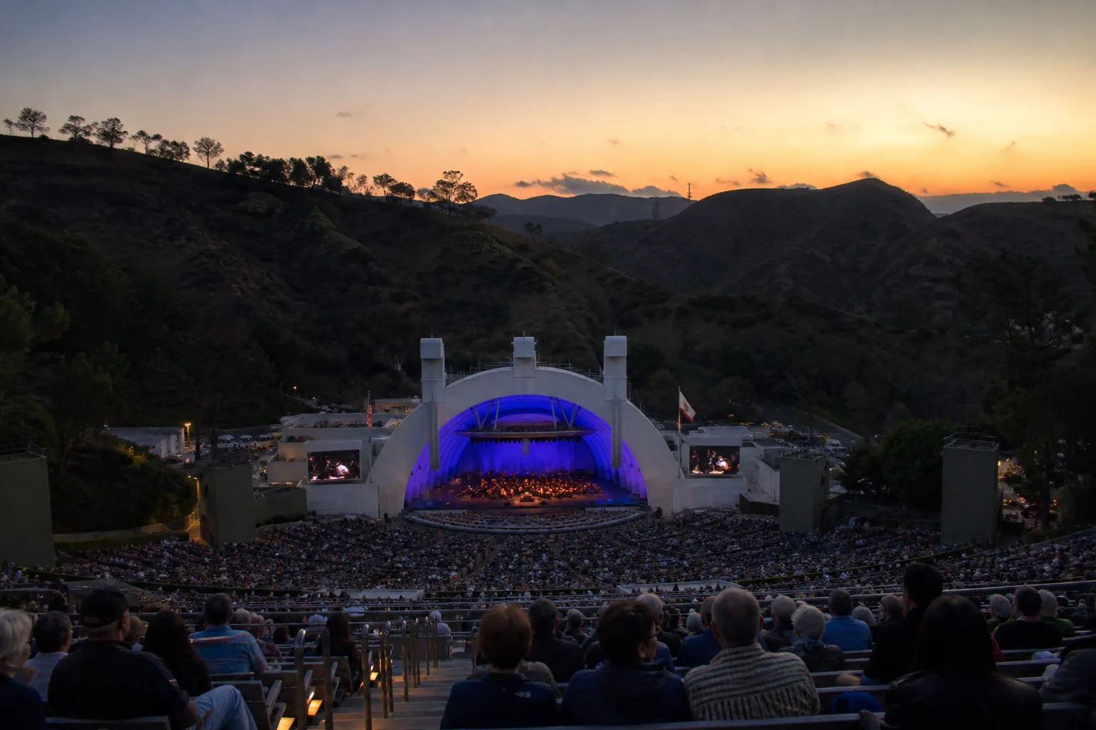 A wide evening landscape shot showing the illuminated white shell of the Hollywood Bowl nestled in the green hills of Los Angeles under a twilight sky.