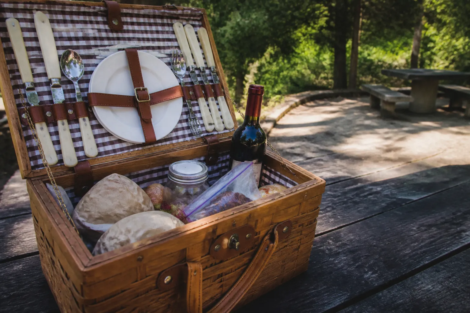 A picnic basket filled with cheeses and fresh bread sitting on a wooden table under the trees.