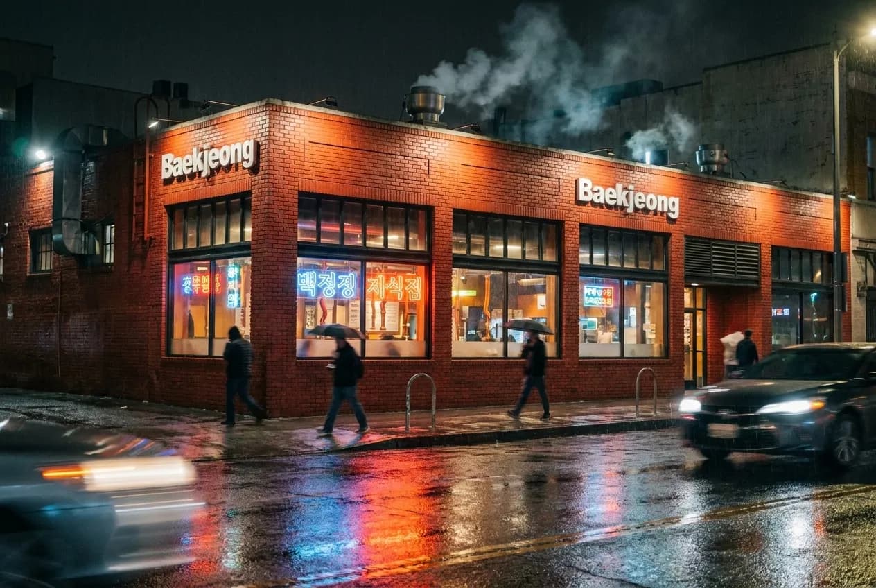 Night exterior of Baekjeong Korean BBQ restaurant on 8th Street in Koreatown, red brick facade with neon signs visible through windows and wet pavement reflecting light.