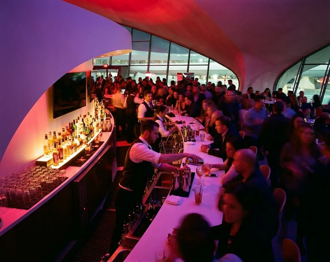 A club bar counter at full tilt with bartenders reaching across a crowded rail and bottles lit from below under deep red and violet lighting