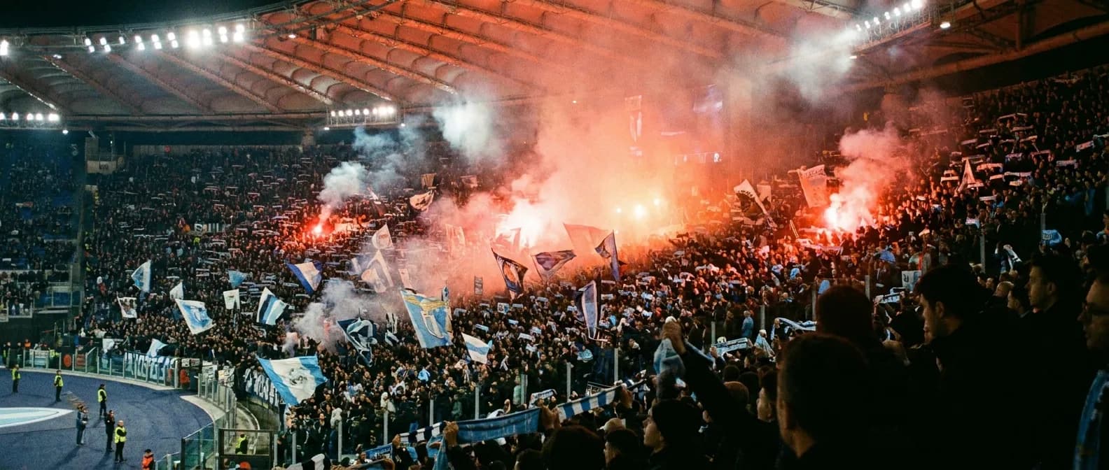 A packed stadium at night with blue and white banners and flares glowing
