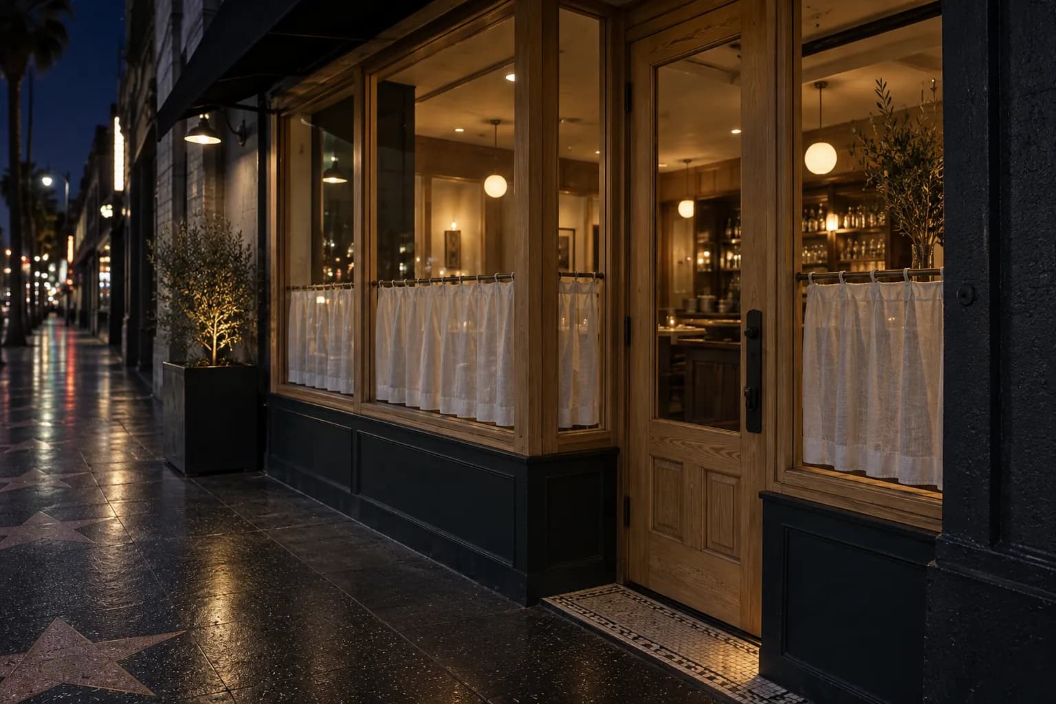 A close night view of a narrow Hollywood Boulevard restaurant frontage, seen diagonally with warm light at the entrance, dark pavement, pale wood details and deep blue street shadows.