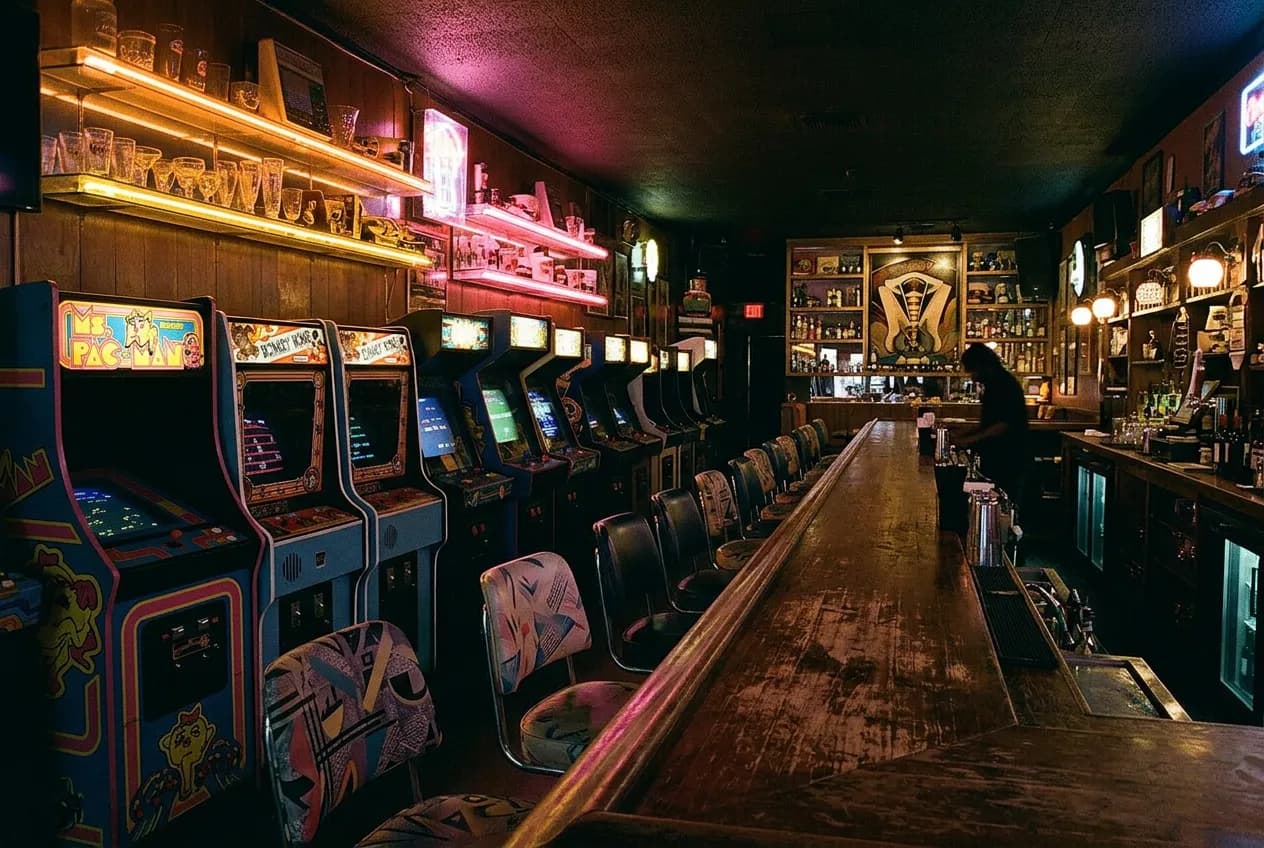 Dim retro Koreatown bar interior with neon light over a scuffed wood counter and arcade cabinets along the wall