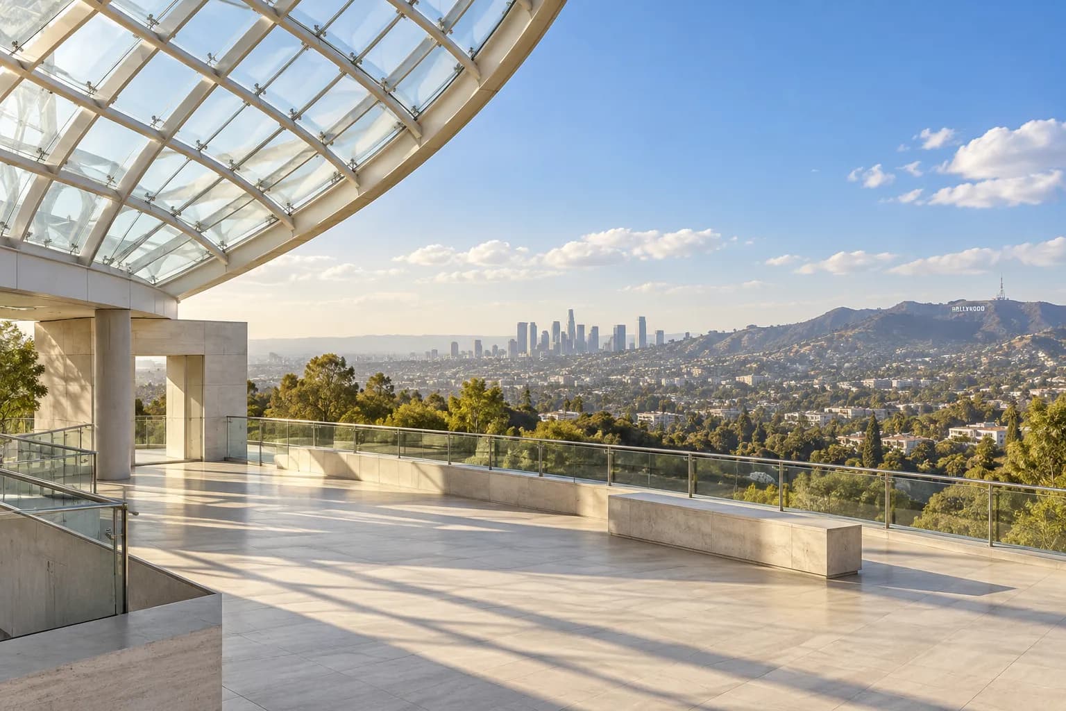 Wide outdoor promenade beneath a curved glass dome, with pale concrete surfaces, low architectural edges and a distant Los Angeles skyline under a clear blue afternoon sky.
