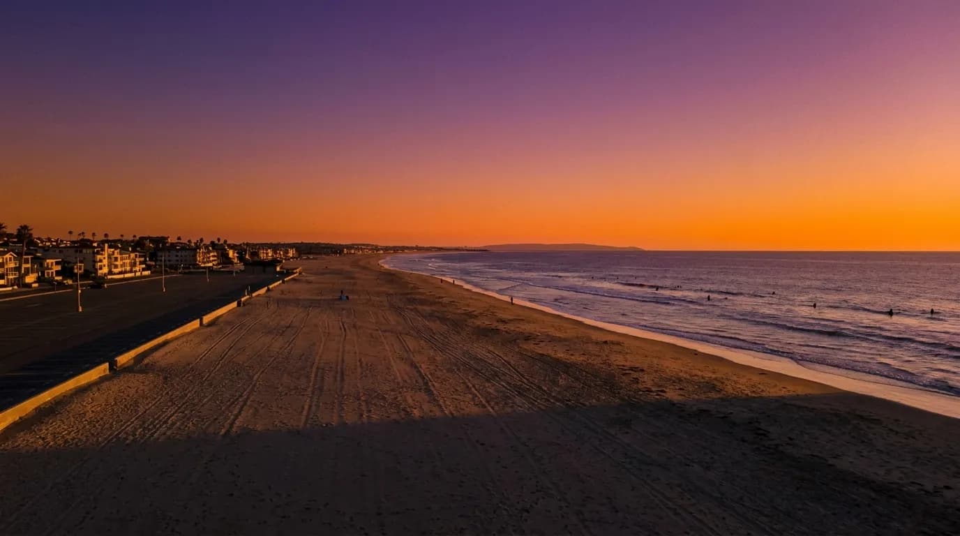 A wide South Bay beach at sunset with surfers in the water and a long parking lot running behind the sand.