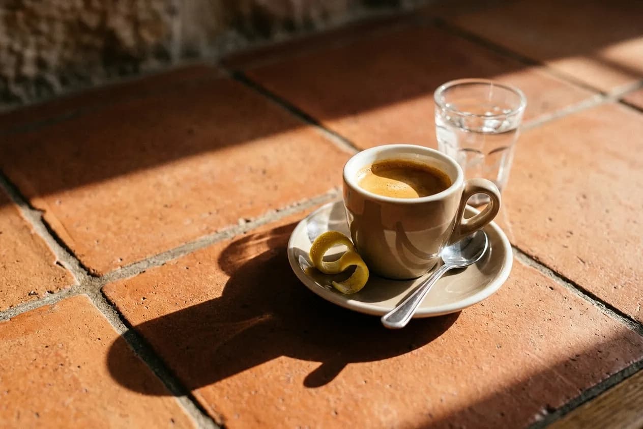 A small ceramic cup of espresso on a saucer on a tiled counter with a glass of water behind, low morning sun casting a hard shadow