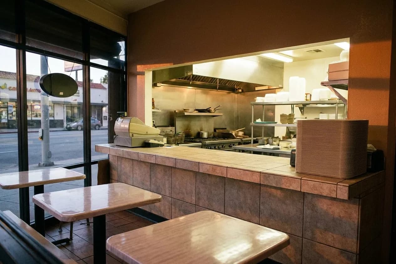 A small tiled order counter at dinner time with a pass-through to a bright kitchen and plastic-topped tables reflected in the window