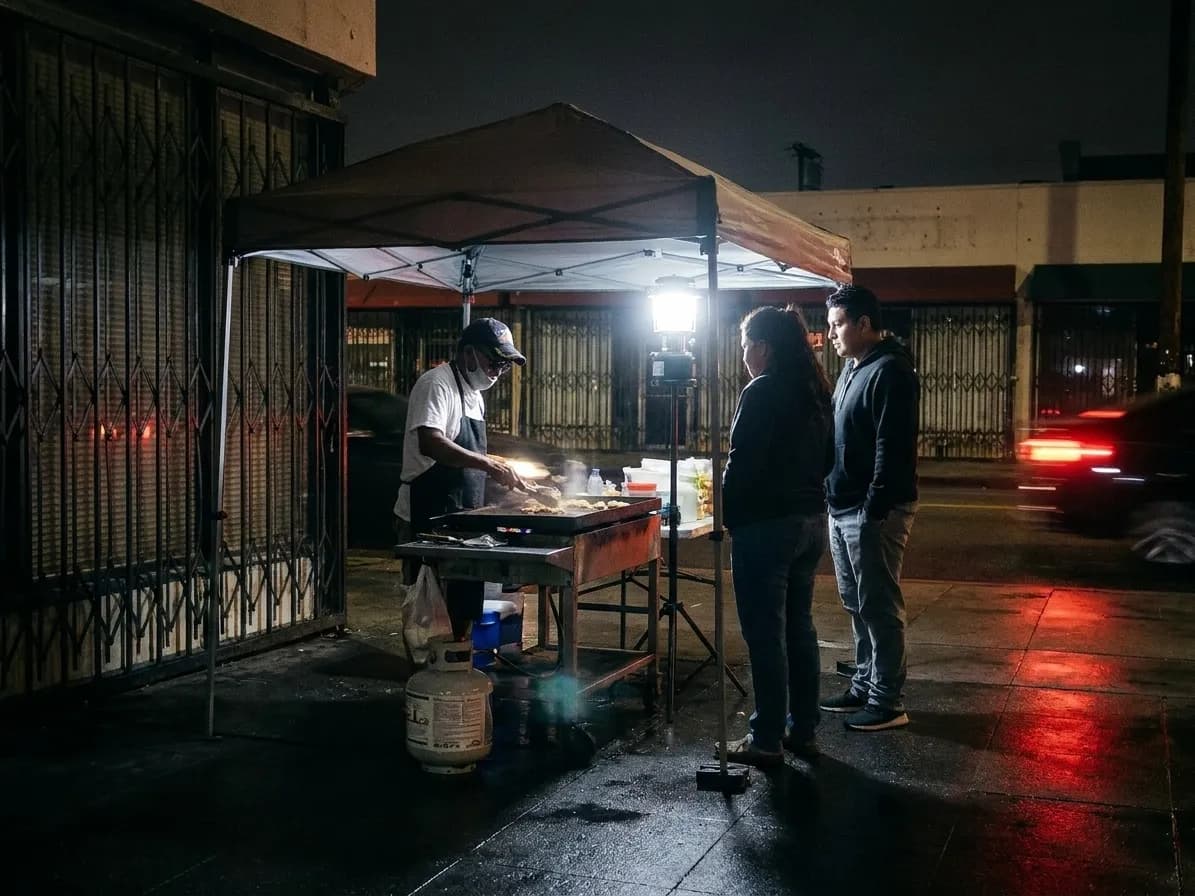 A sidewalk griddle setup under a propane lamp at midnight with a cook and two customers in front of shuttered shopfronts
