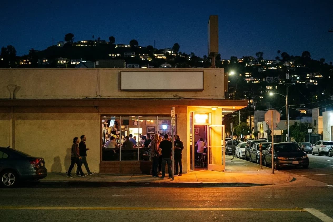 A low neighbourhood bar in Los Angeles at night with warm light spilling from the open door and a hill of house lights behind