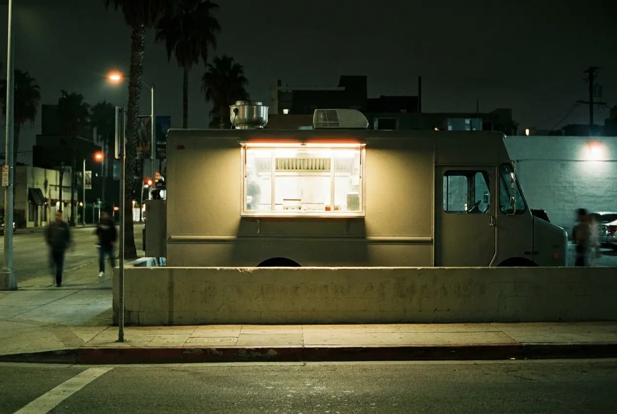A taco truck parked against a low wall, its serving window the brightest thing on the block