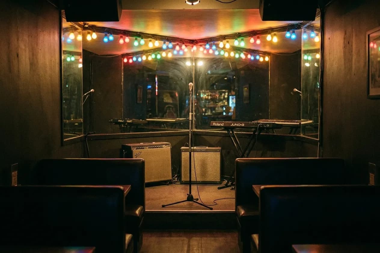 A cramped corner bar stage with amplifiers, a keyboard and a bare mic under a string of coloured bulbs, with dark booths in the foreground