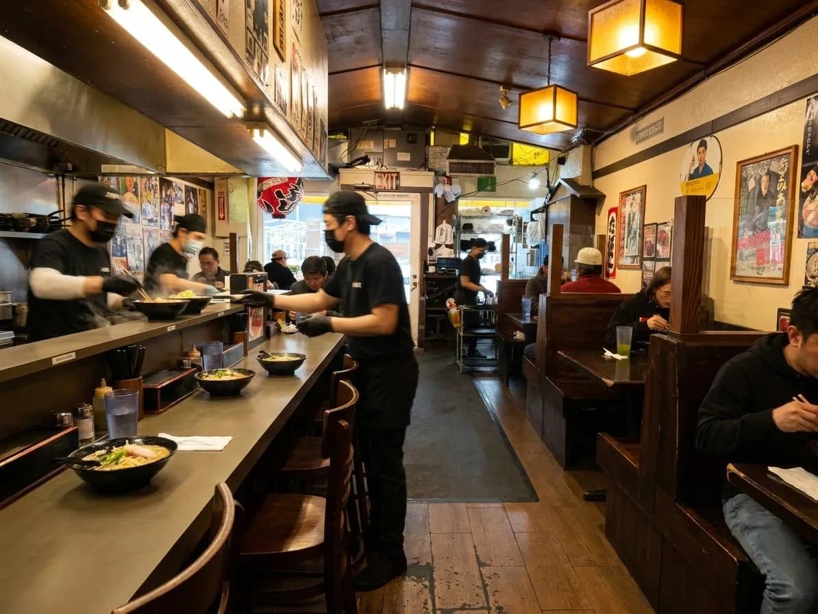 A ramen counter scene with bowls, stools, and warm light