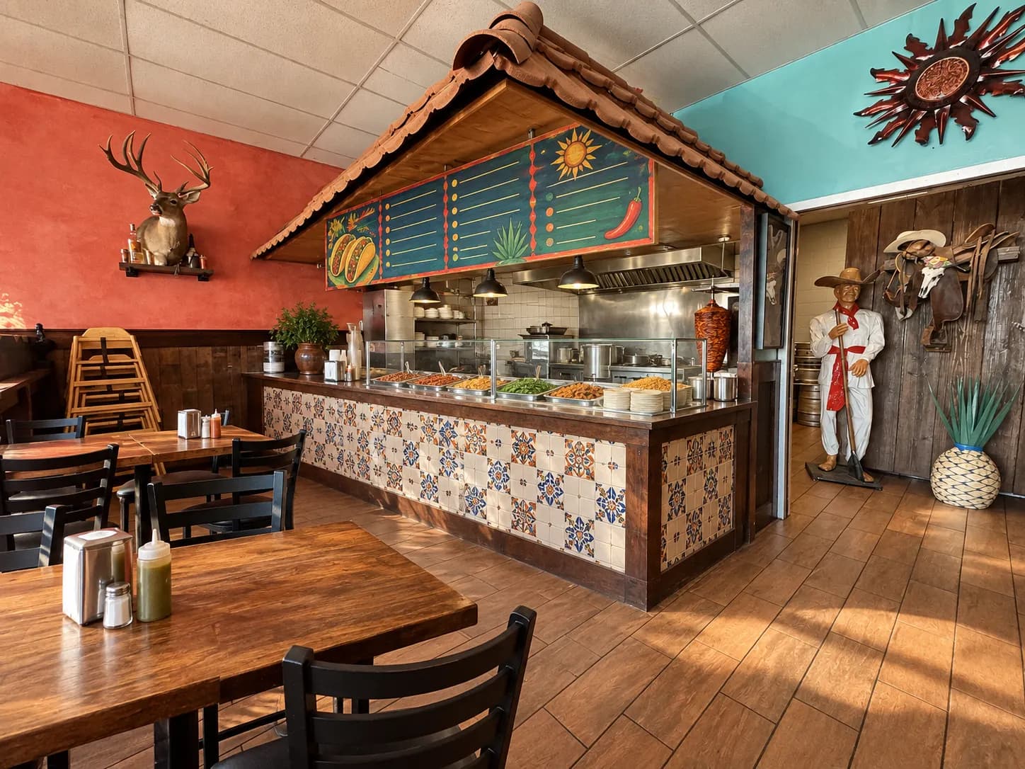 Plain neighbourhood taqueria interior with a tiled order counter, steam trays behind glass, a menu board high on the wall and simple wooden tables in the foreground