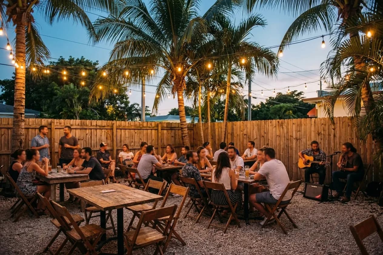 A Miami bar's back garden on a weeknight, mismatched tables on gravel under strings of bulbs with a duo playing acoustic in the far corner