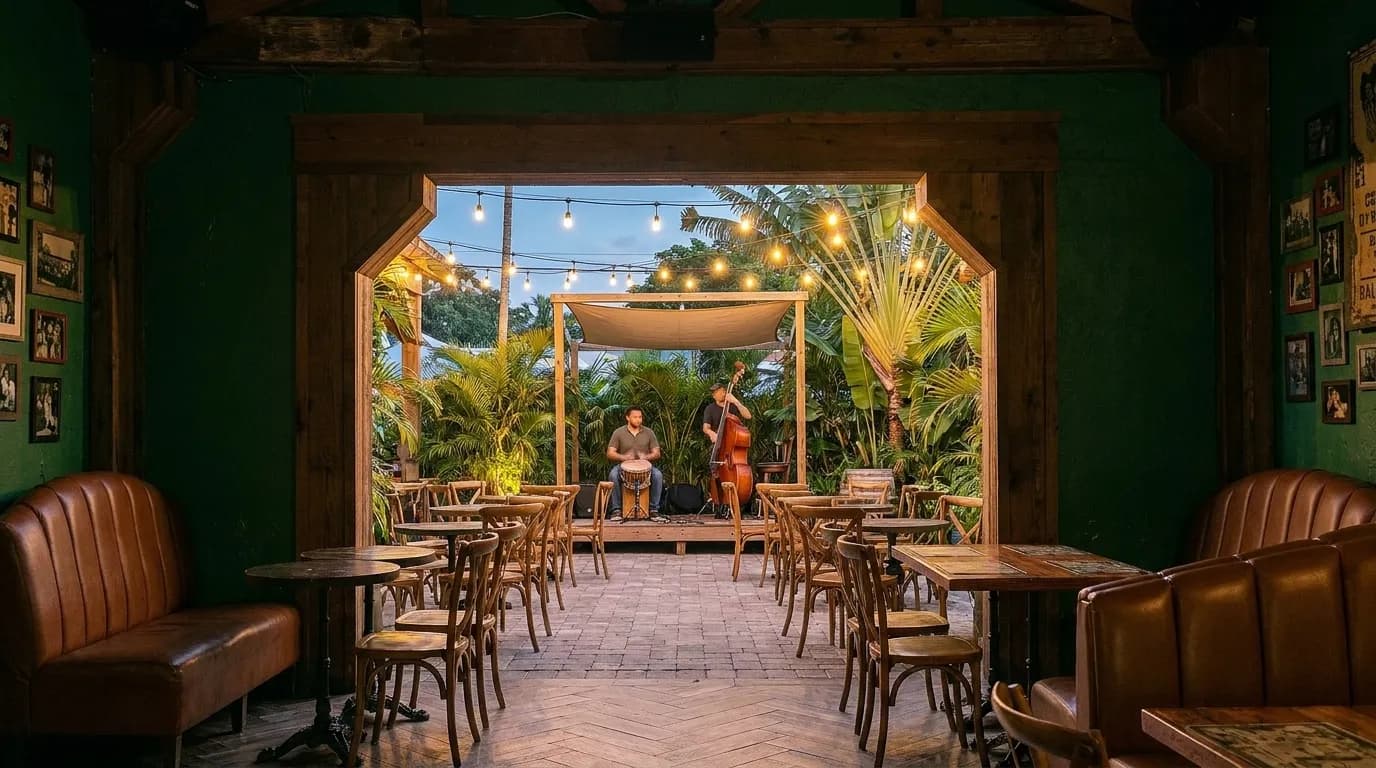 Looking through a timber archway from a dark green bar room into an open-air courtyard at dusk, where a percussionist and an upright bass player set up on a small raised stage under strung bulbs and palms