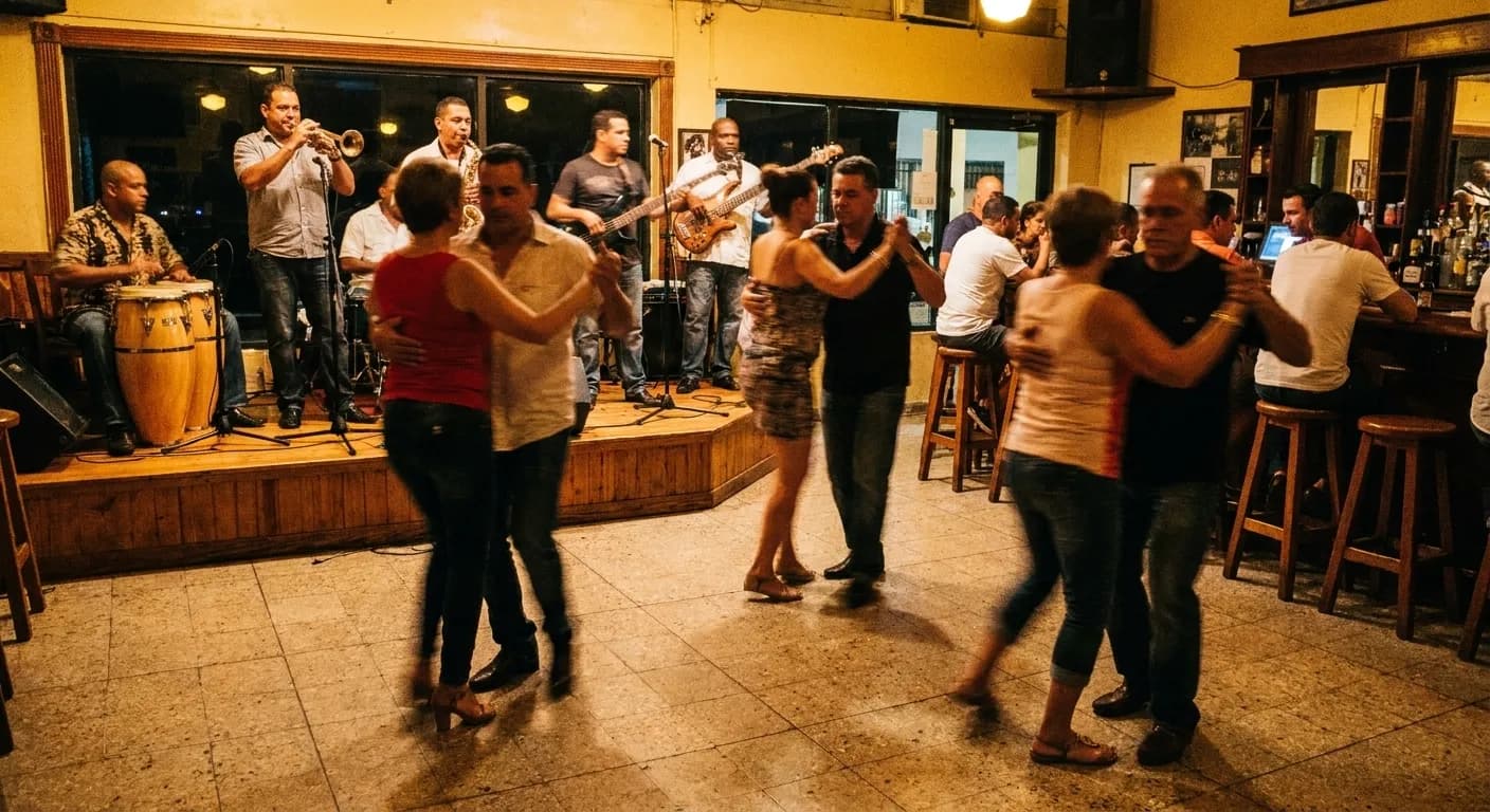 A six-piece Cuban band with congas and trumpet on a shallow stage while four couples dance in motion blur under warm light