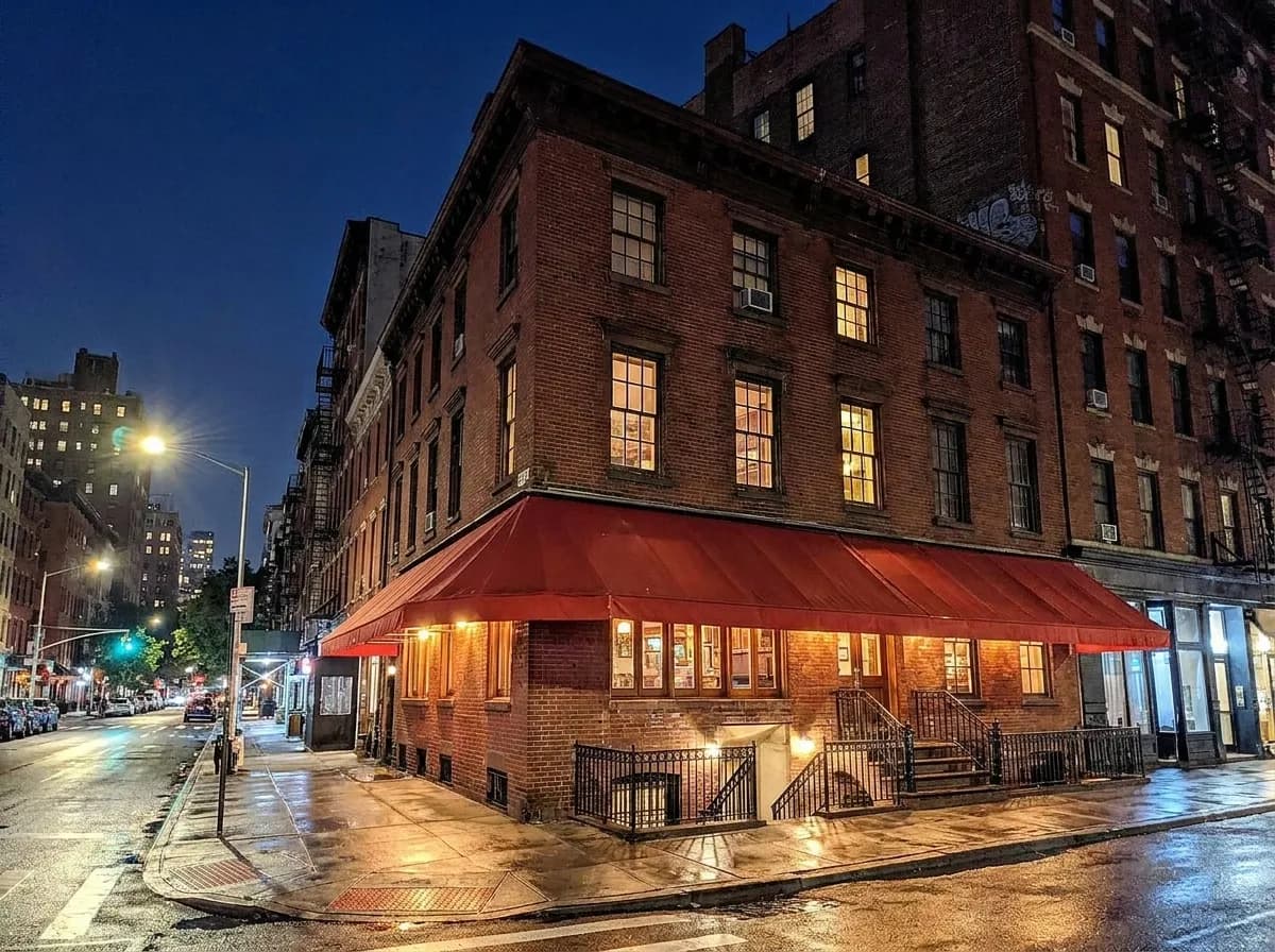 A Greenwich Village corner at night with low brick buildings, a basement stairwell entrance and wet pavement reflecting streetlight