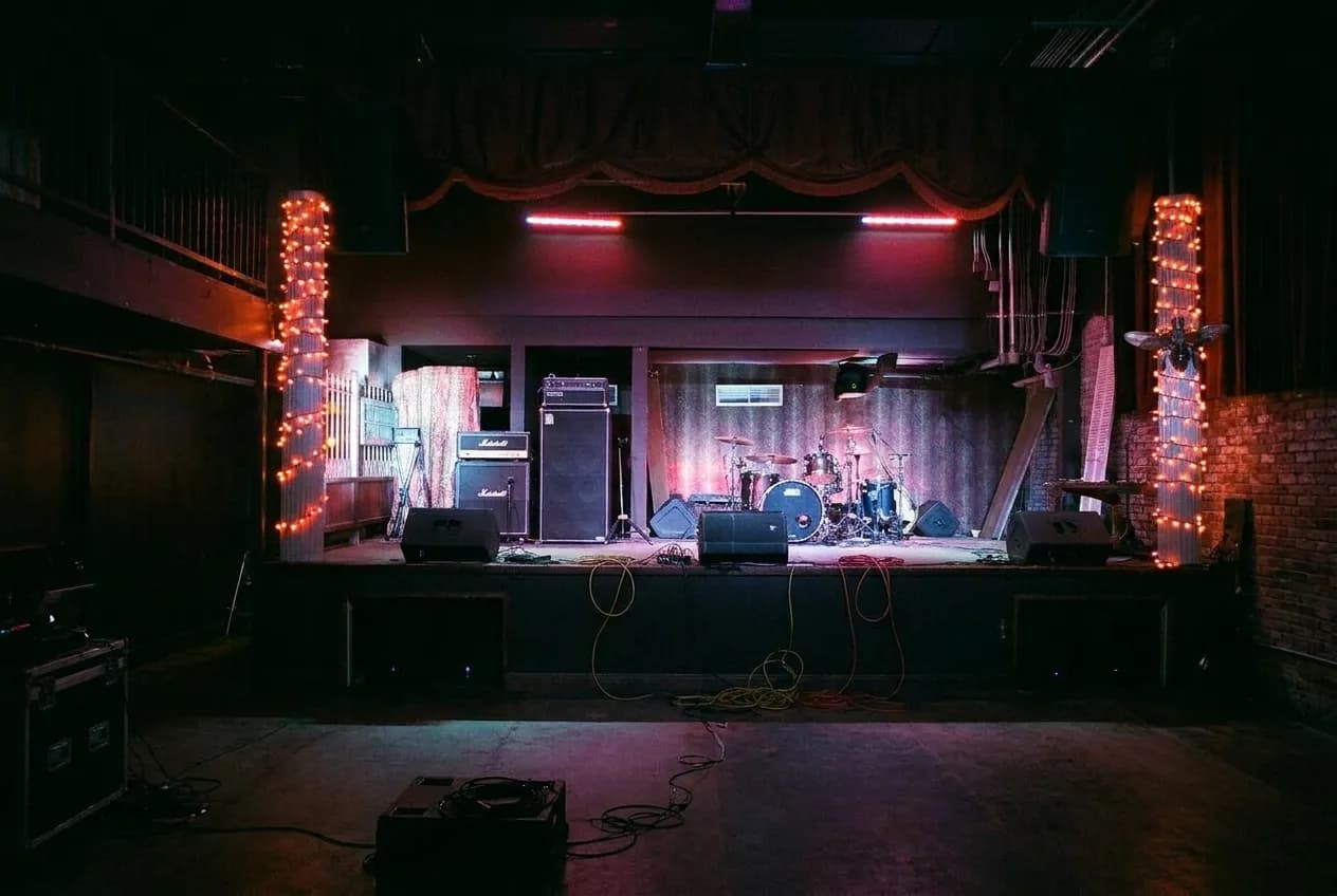 A dark mid-size music venue before doors with a raised stage, amplifier stacks, coiled cables and coloured wash lighting
