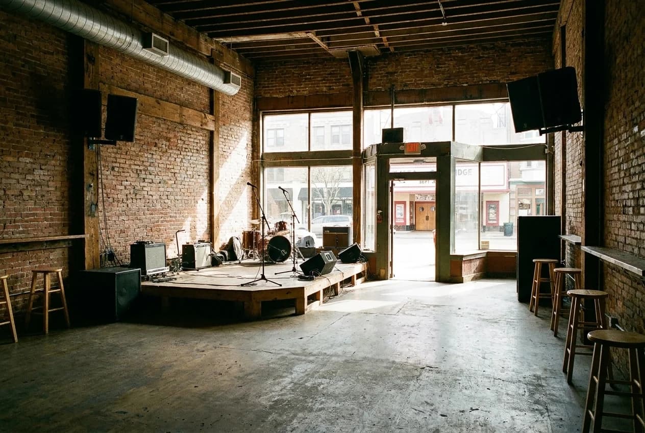 Empty standing-room music venue with brick walls, stage, and microphone stands viewed from the back of the floor.