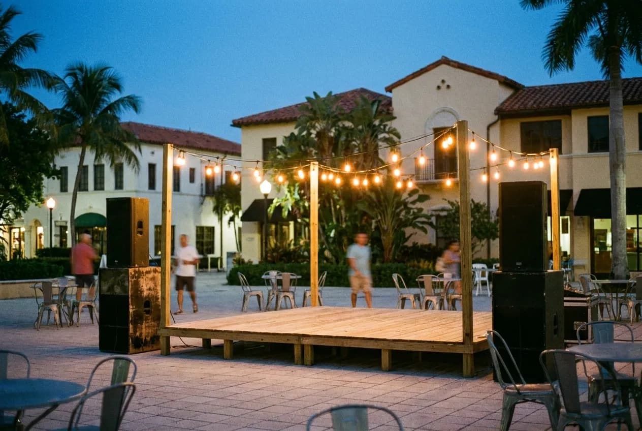 A small outdoor stage under strung lights in the early evening, speaker stacks either side and a paved plaza with scattered chairs in front