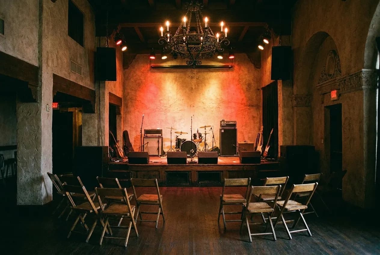 A small low stage in a dim room with instruments set up and coloured lights washing the back wall, a few empty chairs in the foreground