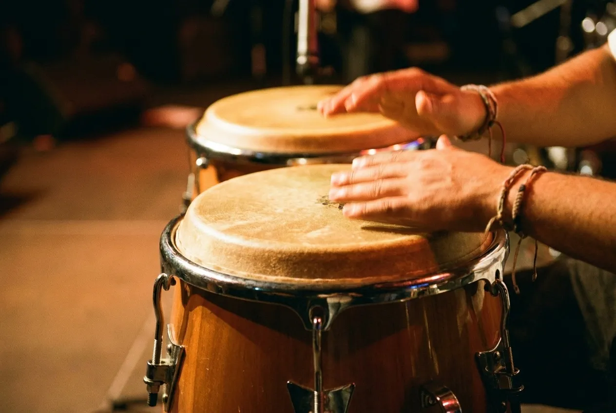 A pair of conga drums photographed close under warm stage light, hands blurred mid-strike on the skin of the nearer drum