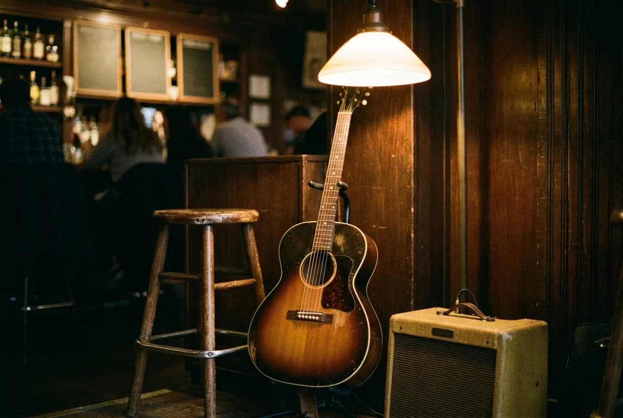 An acoustic guitar propped against a stool in the corner of a bar under a warm hanging lamp, a small amplifier beside it
