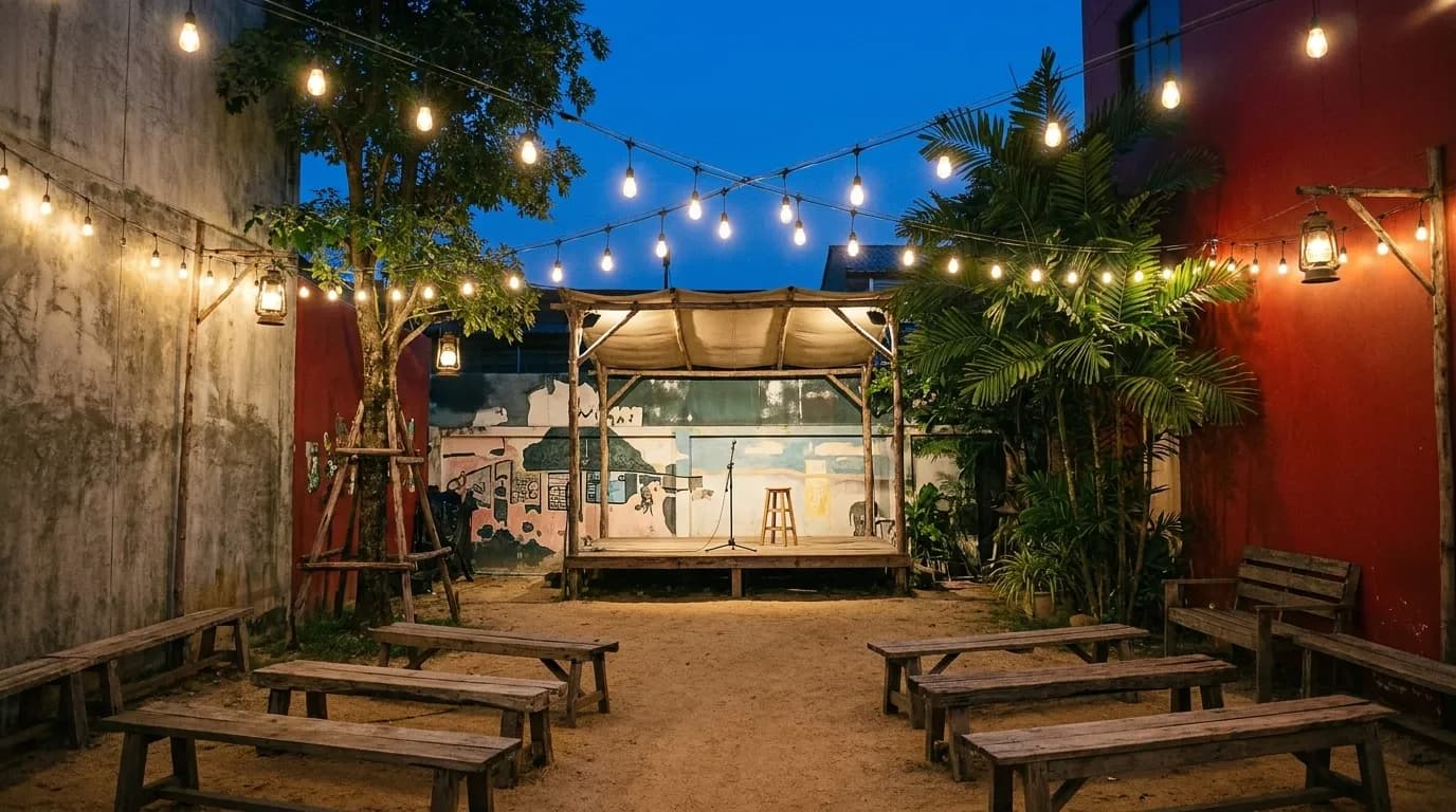 A courtyard music garden strung with bulbs at night, a low covered stage at one end and empty wooden benches arranged loosely in front of it