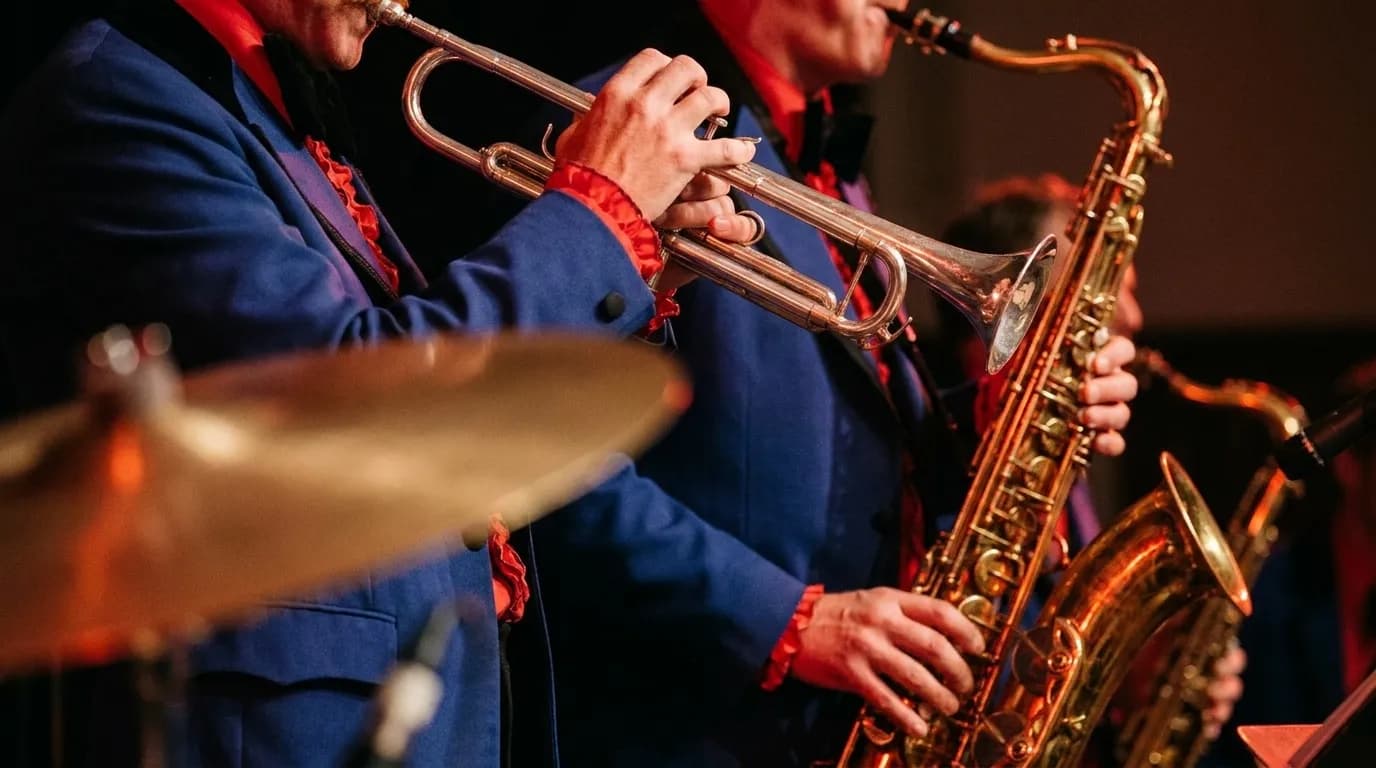 A close view of a brass horn section on a small stage, trumpet and saxophone catching red and amber stage light, a drummer's cymbal blurred in the foreground