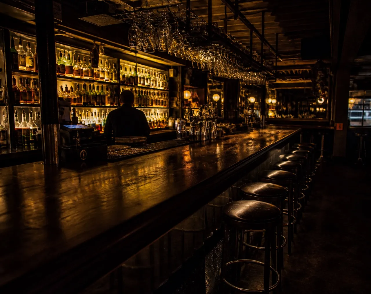 Dark bar interior with a lit back bar and stools along the counter