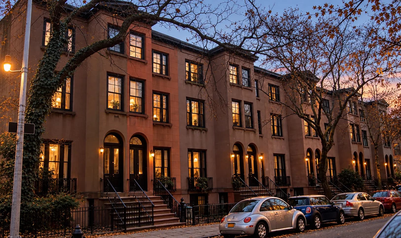 Quiet Brooklyn residential street at dusk with rows of brownstone stoops, mature street trees and warm light in ground-floor windows