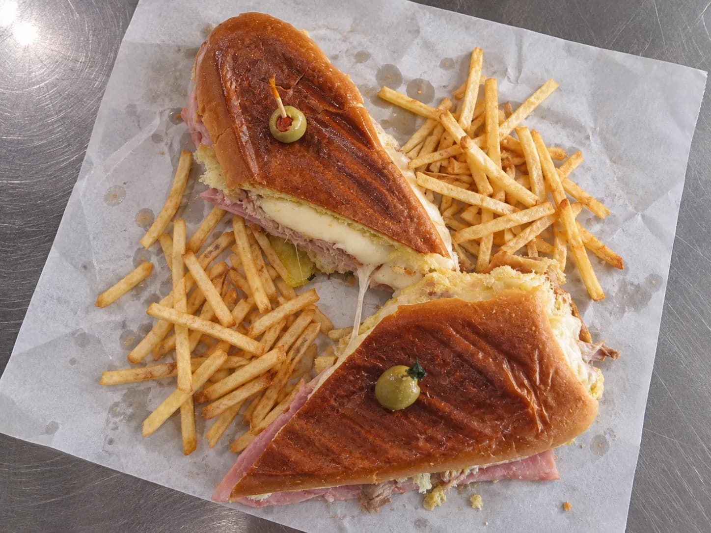 Pressed Cuban sandwich cut on the diagonal on greaseproof paper, flattened crust with dark griddle marks and filling visible at the cut faces