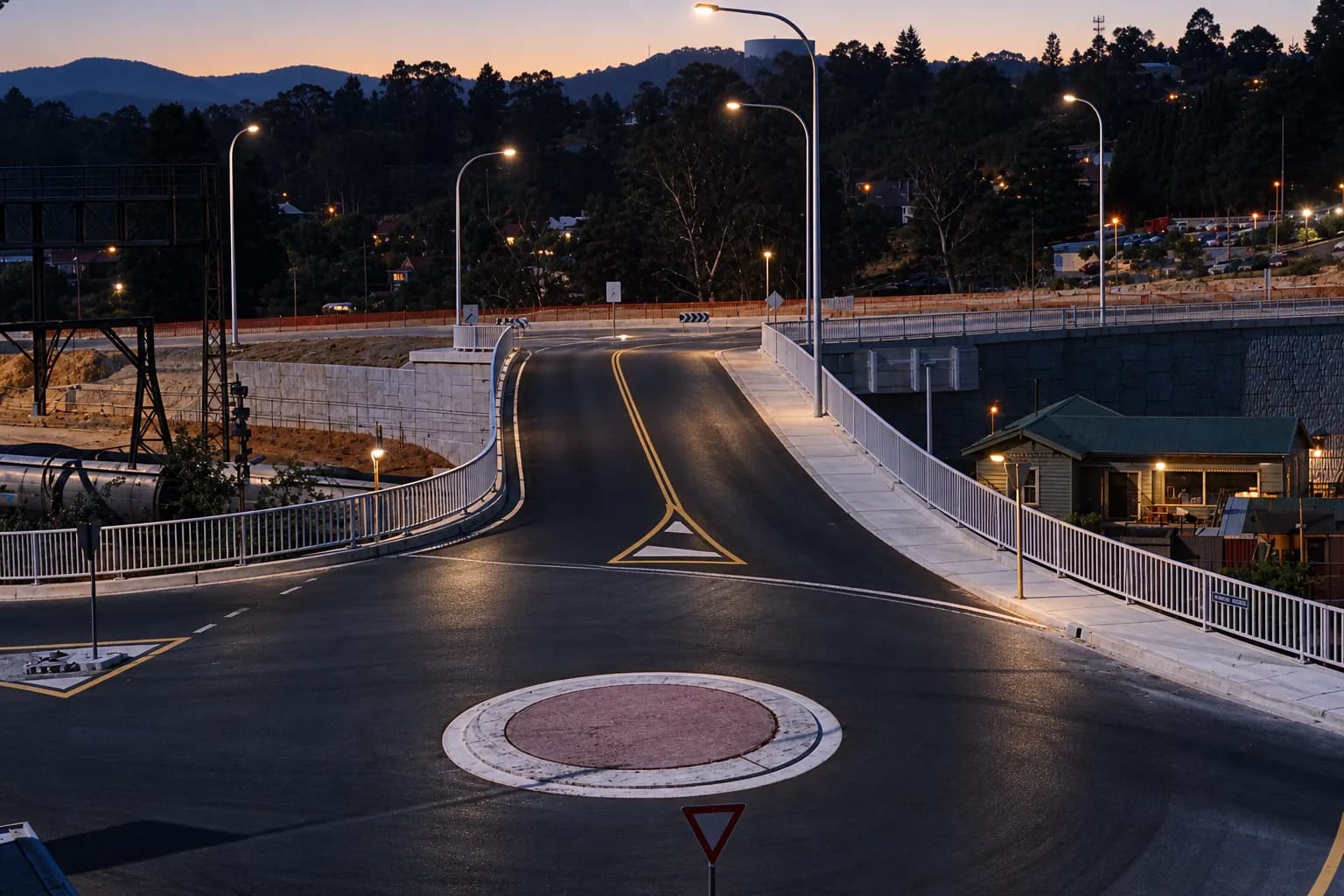 Wide arterial road at dawn feeding toward a freeway on-ramp, with overhead sign gantries silhouetted and pale sky brightening behind hills