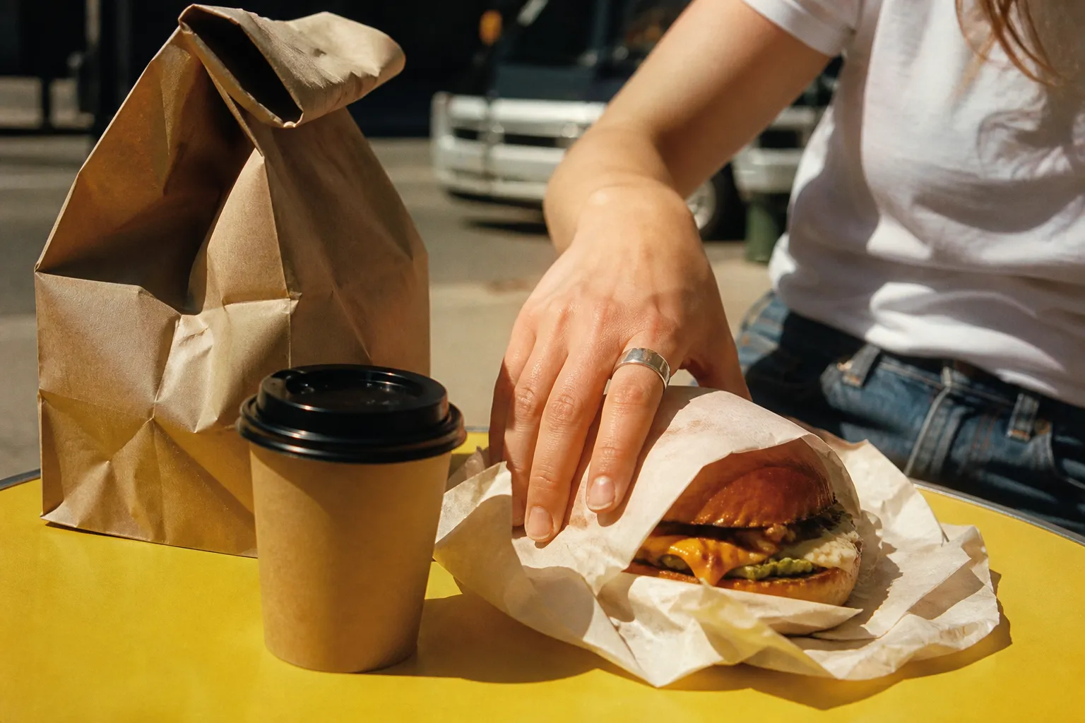 Casual outdoor table with a paper takeout bag, coffee cup and a wrapped burger in warm afternoon light