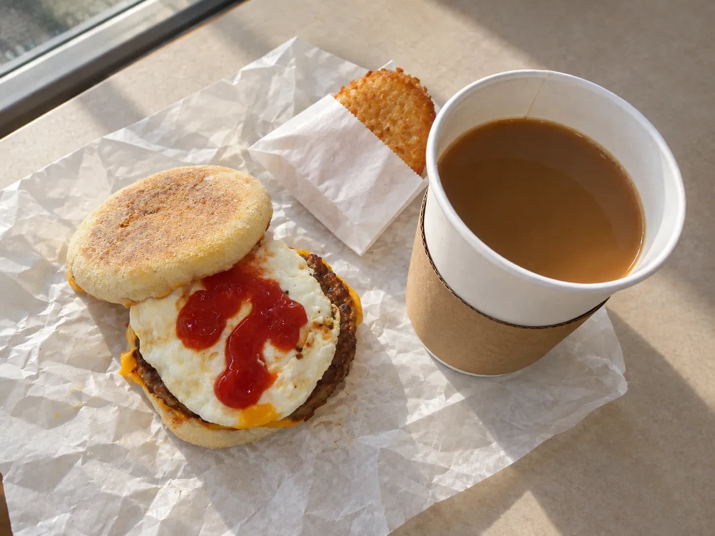Toasted English muffin breakfast sandwich with a round egg and sausage patty on its unfolded paper wrapper, beside a paper coffee cup and a hash brown