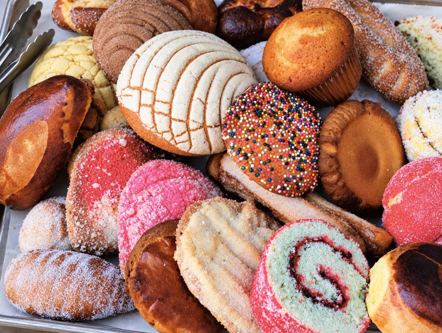Bakery tray of pan dulce with conchas in pink, yellow and white showing crackled sugar shells, alongside other sweet breads
