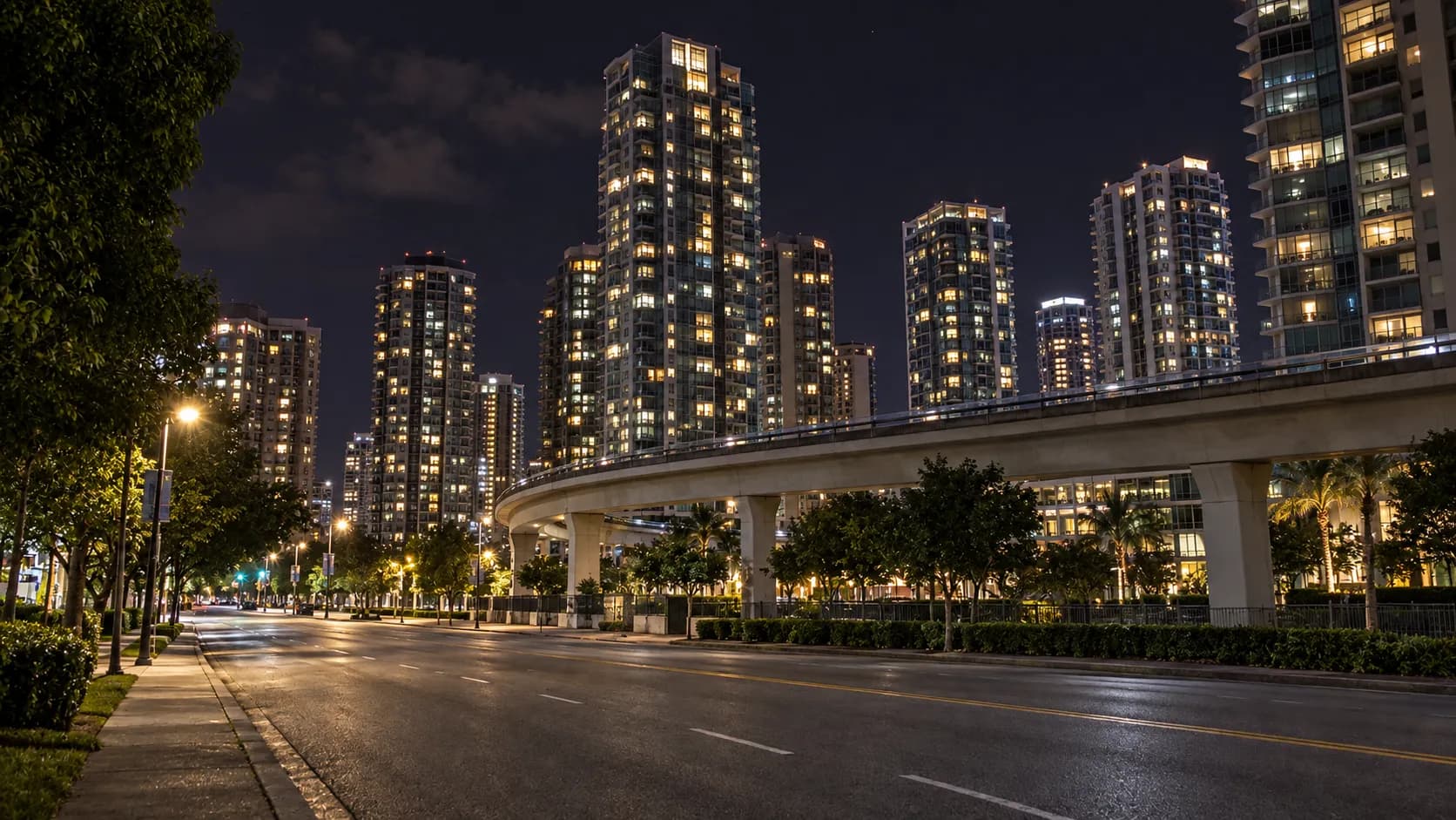Dense cluster of residential high-rise towers at night with unevenly lit glass facades and an elevated transit guideway curving in the middle distance