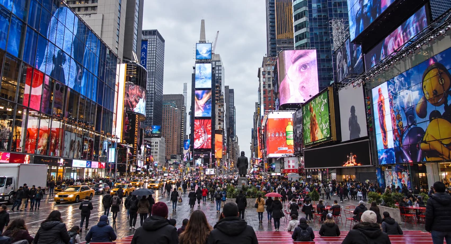 Busy pedestrian plaza between converging avenues with tall buildings clad in illuminated screens on every side and red bleacher steps