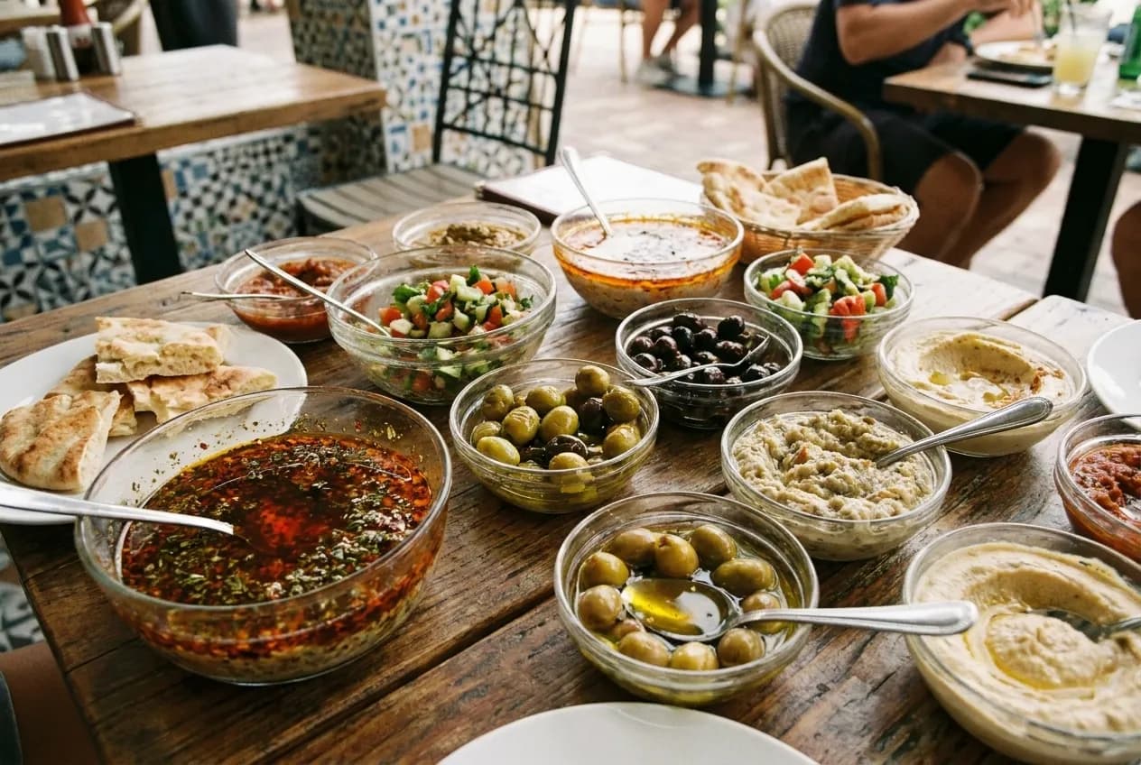 A table seen from above crowded with small dishes of dips, olives, salads and flatbread, the surfaces catching soft directional light