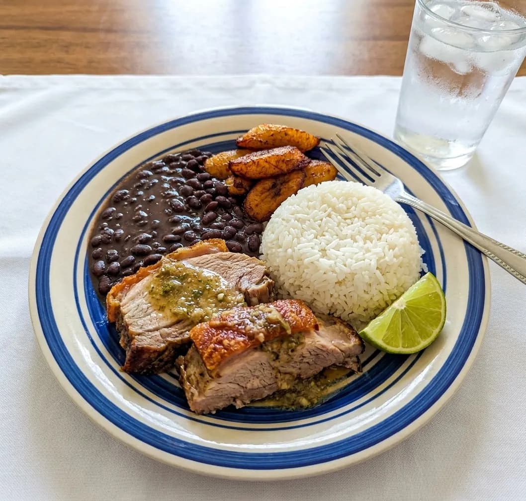 A plate of Cuban roast pork with white rice, black beans, fried sweet plantains and a wedge of lime on a white tablecloth