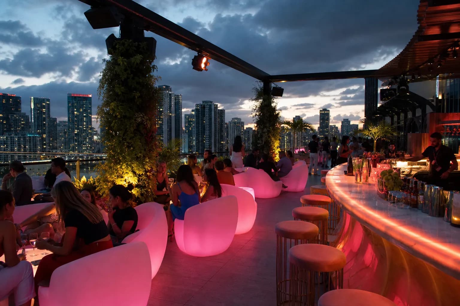 A stylish rooftop lounge illuminated by pink lights with guests enjoying cocktails overlooking the Miami city skyline at dusk.