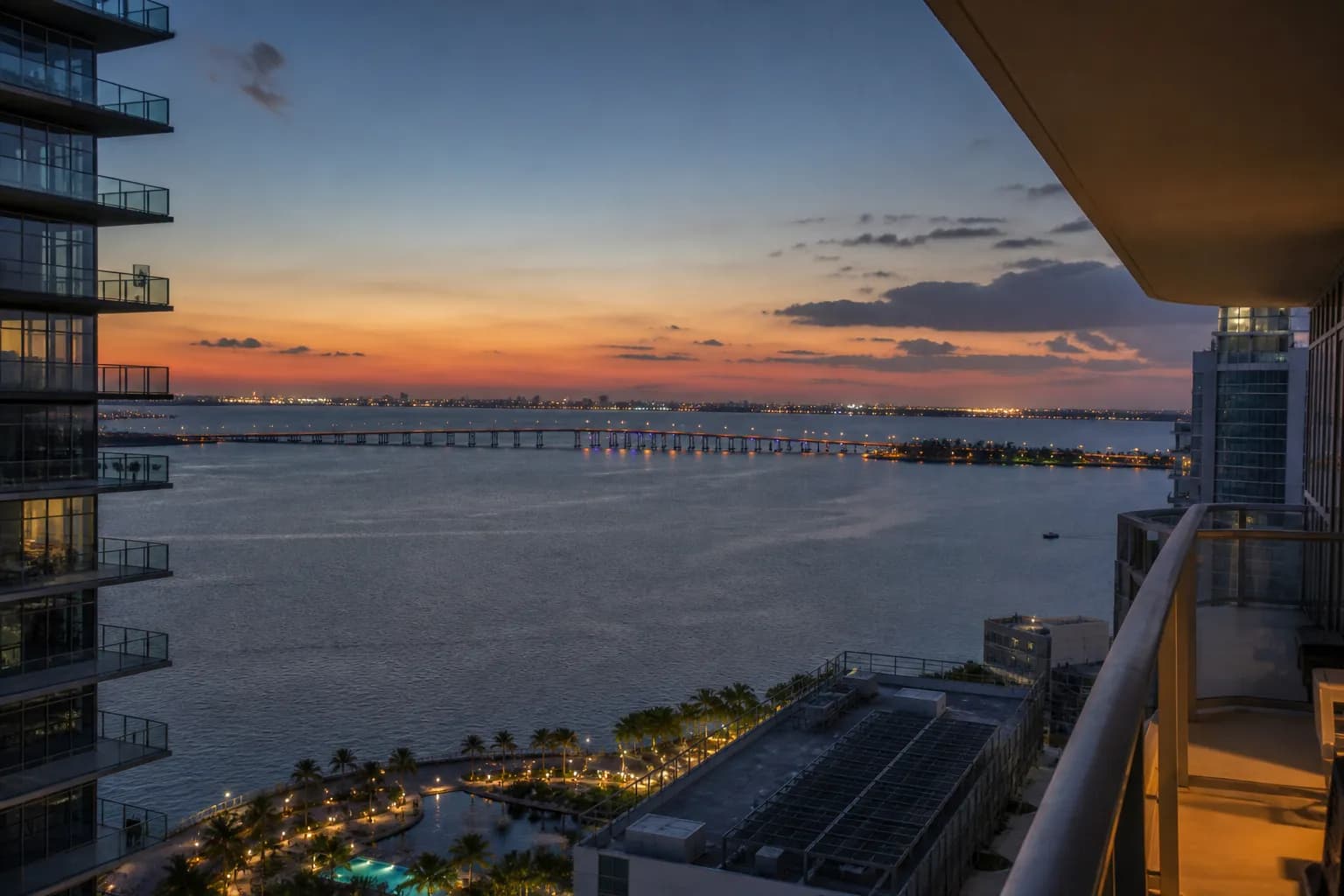 A panoramic view from a high-rise balcony looking over Biscayne Bay and distant bridge lights during the blue hour in Miami.