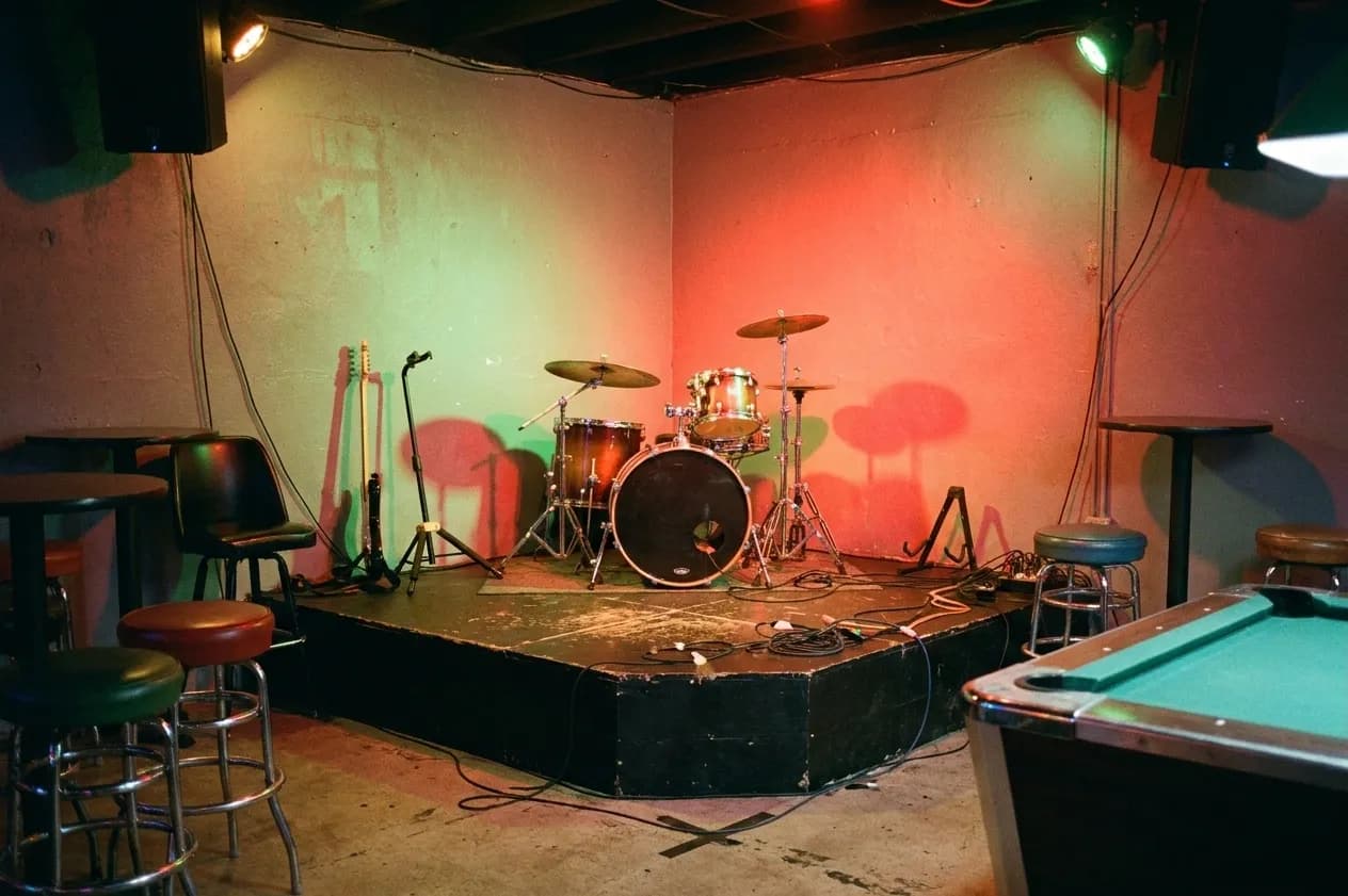 The corner stage of a small Miami neighborhood music bar between sets, with a drum kit and guitar stands left standing under red and green stage light, cables taped to the floor and the edge of a pool table in the foreground