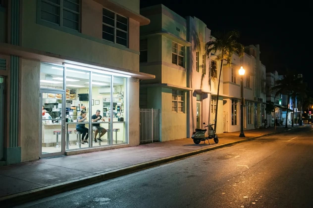 A South Beach side street at three in the morning, a small brightly lit sandwich counter seen through plate glass with two people eating at the window bar, a scooter at the kerb and pastel Art Deco facades beyond