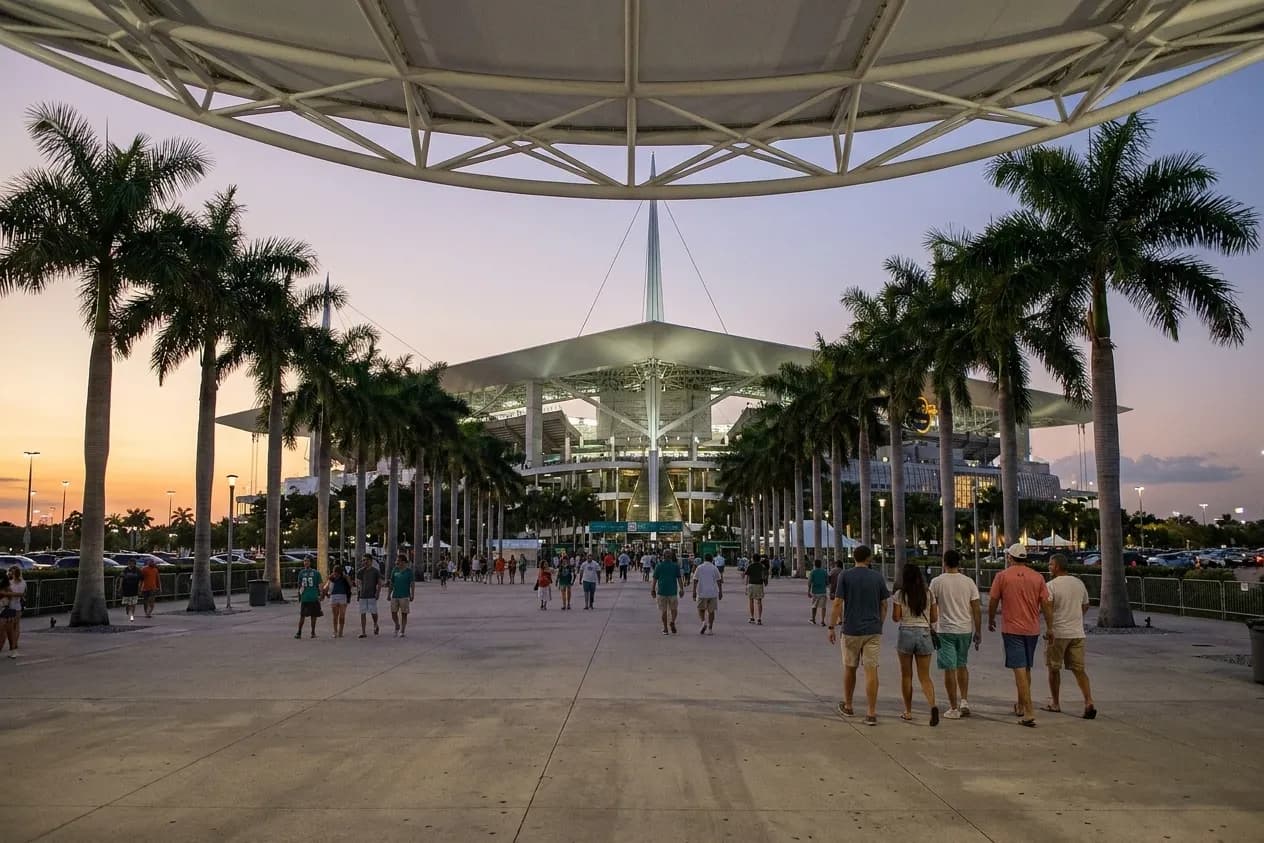The approach plaza of a large Miami stadium an hour before an evening event, palms and a floodlit white steel canopy above small groups walking to the gates under an orange dusk sky