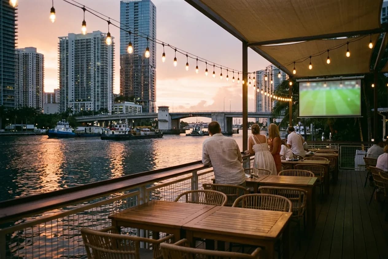 A waterfront terrace at dusk with low tables, string lights overhead and a wide dark river behind, a screen glowing at the far end of the deck
