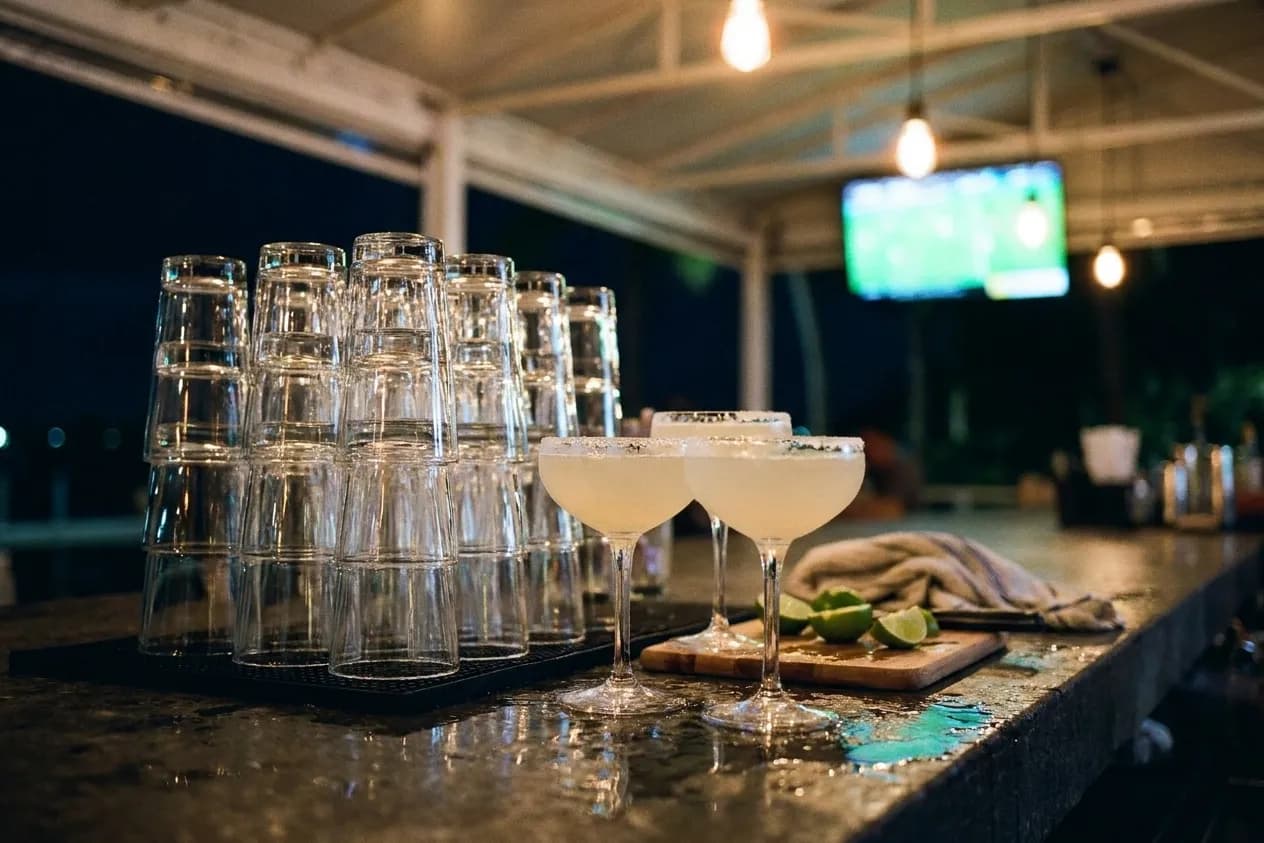 A bartender's rail at night with three margaritas in coupe glasses, a lime wedge board and the blur of a covered patio behind