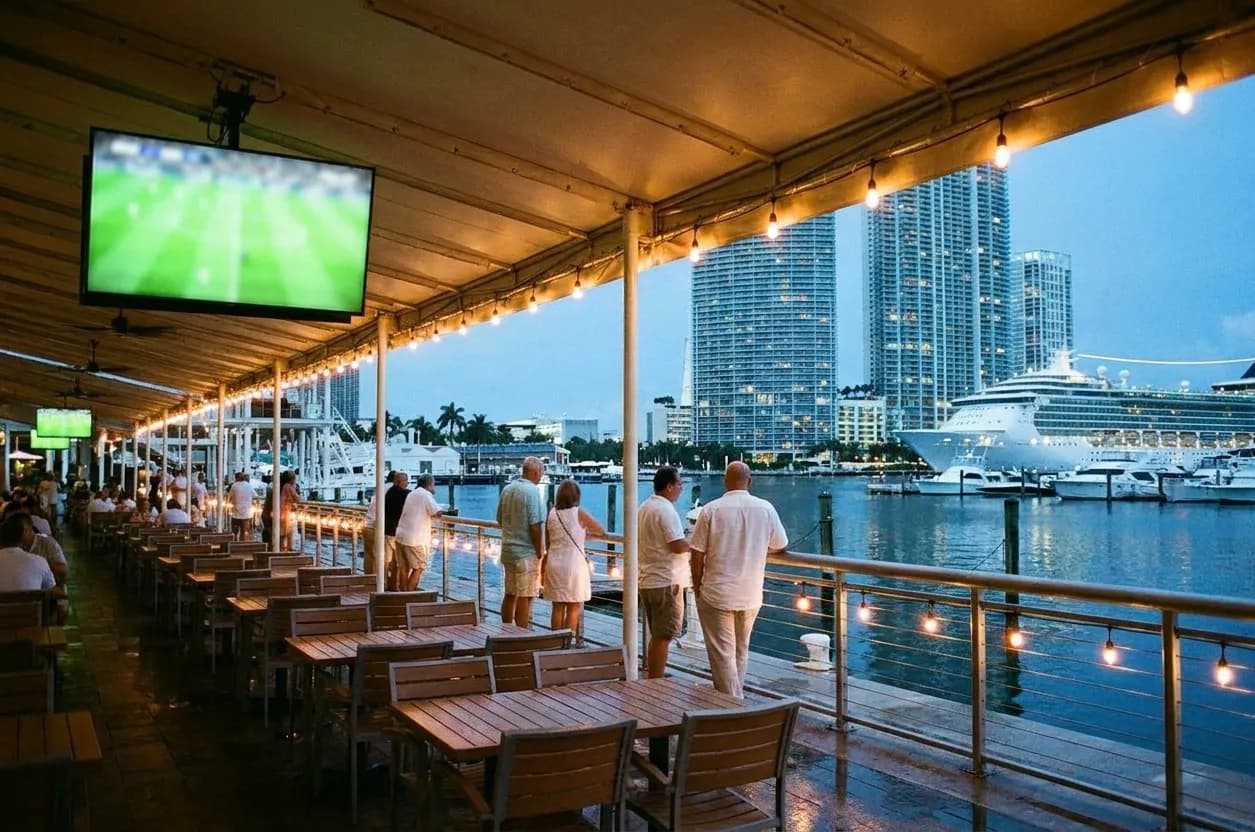 A wide open-air waterfront dining terrace in downtown Miami at dusk, cruise ship lights beyond the rail and a large screen glowing under an awning