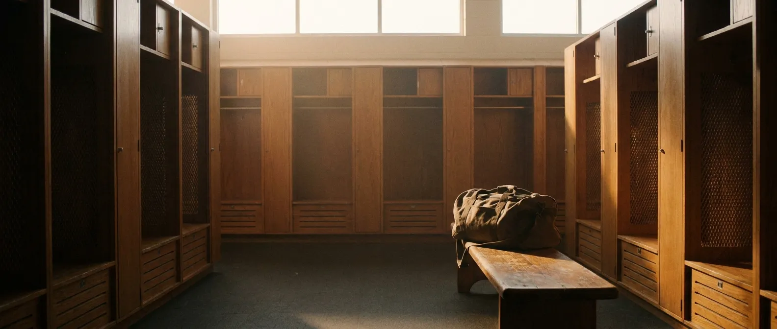 Rows of empty lockers in a football facility at dawn with a single duffel bag on a wooden bench